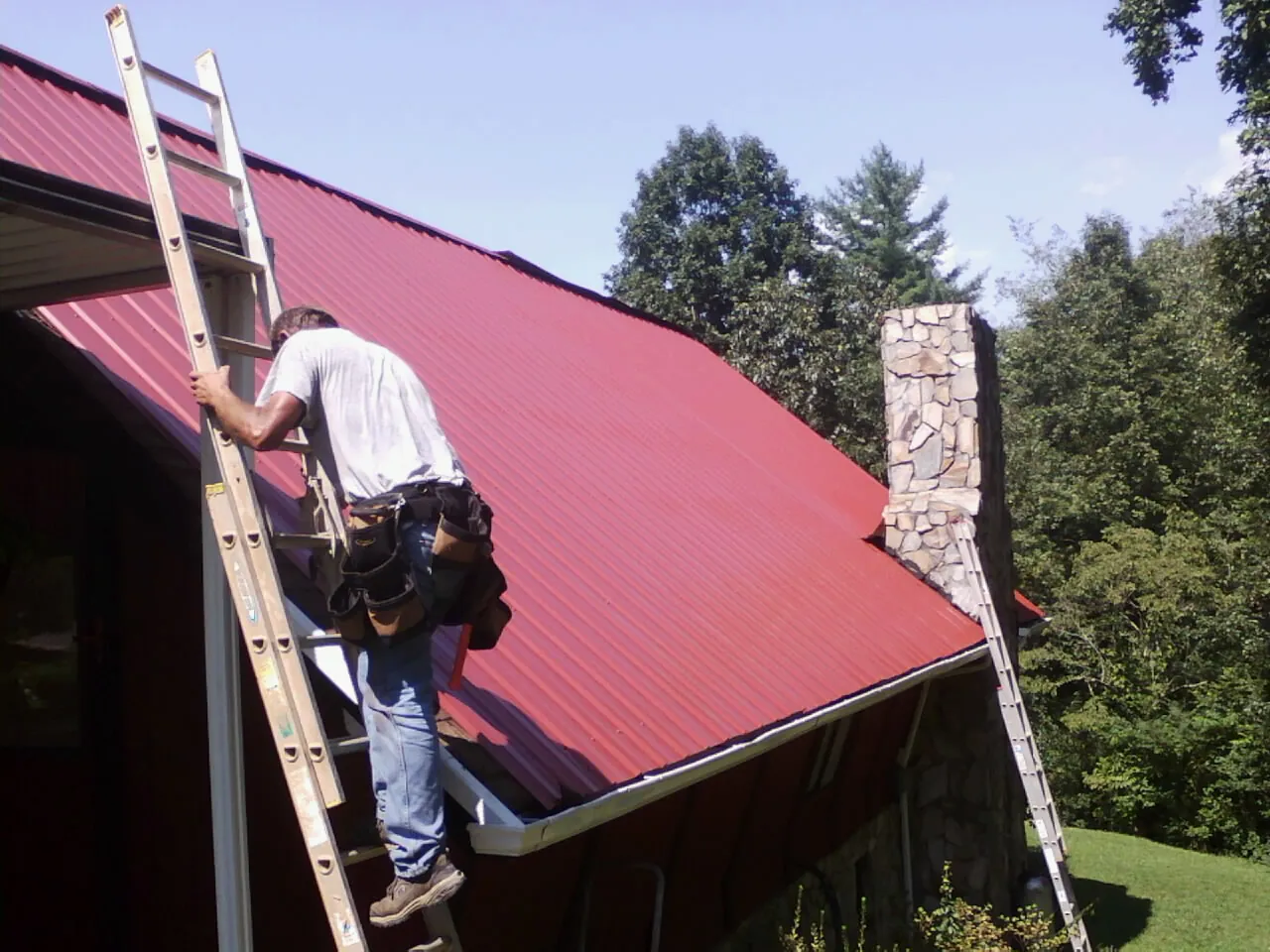 Roofer on a ladder beside a red metal roof and stone chimney — Soffit & Fascia Repair by Roofing Brothers in Chemung